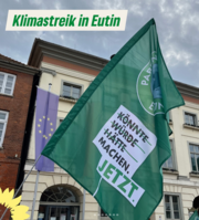 Grünes Banner bei einer Demonstration in Eutin vor dem Rathaus. Auf der Fahne steht: „Könnte, würde, hätte – MACHEN. JETZT.“ Im Hintergrund weht eine Europaflagge an einem alten Gebäude. Darüber eingeblendet der Schriftzug „Klimastreik in Eutin“