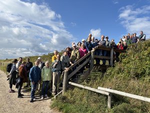 Eine große Gruppe von Menschen steht bei sonnigem Wetter auf einem Holzsteg und daneben auf einem Sandweg inmitten von Dünen und Büschen. Die Gruppe schaut in Richtung Kamera und wirkt wie bei einem gemeinsamen Ausflug oder einer Führung in der Natur. Über ihnen blauer Himmel mit weißen Wolken.